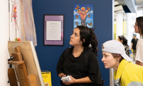Two young women closely observe artwork displayed on a wall in an indoor setting, with other people and art visible in the background.