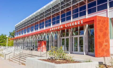 Modern library exterior with glass facade and bold red accents labeled "Auraria Library." Sunny day with stairs and sunflowers leading to entrance.