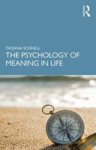 A book cover featuring a blue background with the title "The Psychology of Meaning in Life" by Tatjana Schnell. Foreground shows a compass on a sandy beach.