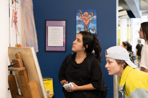 Auraria Creates event spotlights campus scholars and creatives Two young women closely observe artwork displayed on a wall in an indoor setting, with other people and art visible in the background.