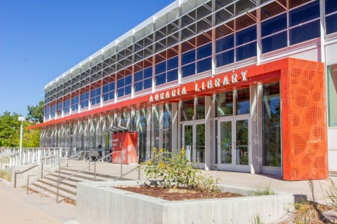 New Badge-Required Hours for the Auraria Library Modern library exterior with glass facade and bold red accents labeled "Auraria Library." Sunny day with stairs and sunflowers leading to entrance.