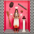 A woman in traditional attire stands against a pink background. She's flanked by cultural artifacts, pine cones on a pedestal, conveying a ceremonial tone.